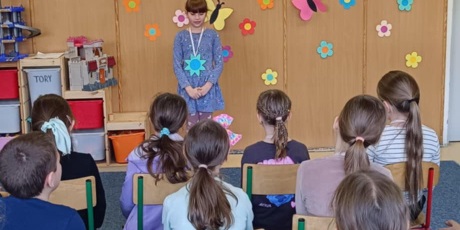 Powiększ grafikę: Grupa dzieci siedzi tyłem.  Dziewczynka stoi przodem i recytuje wiersz.