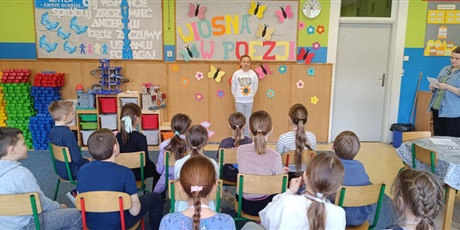Powiększ grafikę: Grupa dzieci siedzi tyłem.  Dziewczynka stoi przodem i recytuje wiersz.