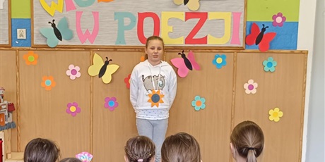 Powiększ grafikę: Grupa dzieci siedzi tyłem.  Dziewczynka stoi przodem i recytuje wiersz.
