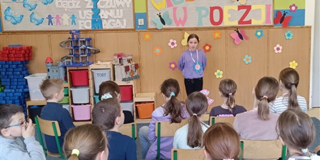 Powiększ grafikę: Grupa dzieci siedzi tyłem.  Dziewczynka stoi przodem i recytuje wiersz.