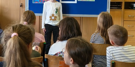 Powiększ grafikę: Chłopiec w białej bluzce stoi zwrócony przodem do publiczności.  Za nim stoi granatowa tablica z kolorową dekoracją i napisem II Edycja Konkursu Recytatorskiego „W świecie Marii Konopnickiej”.