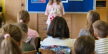Powiększ grafikę: Dziewczynka w różowej bluzce stoi zwrócona przodem do publiczności.  Za nią stoi granatowa tablica z kolorową dekoracją i napisem II Edycja Konkursu Recytatorskiego „W świecie Marii Konopnickiej”.