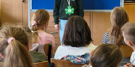 Powiększ grafikę: Dziewczynka w ciemnej bluzie stoi zwrócona przodem do publiczności.  Za nią stoi granatowa tablica z kolorową dekoracją i napisem II Edycja Konkursu Recytatorskiego „W świecie Marii Konopnickiej”.