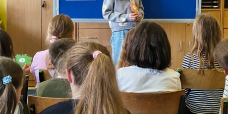 Powiększ grafikę: Dziewczynka w szarej bluzie stoi zwrócona przodem do publiczności.  Za nią stoi granatowa tablica z kolorową dekoracją i napisem II Edycja Konkursu Recytatorskiego „W świecie Marii Konopnickiej”.