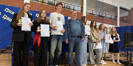 Powiększ grafikę: Dzieci w wieku 13-14 lat stoją i pokazują dyplomy. Obok stoi mężczyzna. Wszyscy są uśmiechnięci.