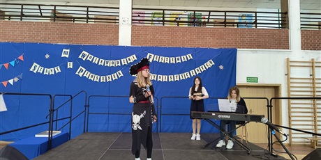 Powiększ grafikę: Uczennica w wieku ok 12 lat przebrana za piratkę trzyma mikrofon. W tle siedzi przy pianinie kobieta oraz stoi dziewczynka w wieku ok 12 lat.