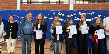 Powiększ grafikę: Grupa uczniów ok 13 lat i nauczycieli stoi uśmiechnięta. Dzieci mają w rękach kartki z dyplomami. W tle dekoracja.