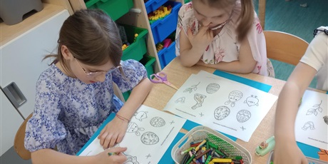 Powiększ grafikę: Dzieci wykonują pracę plastyczną "W wielkanocnym koszyczku" - dziewczynka w sukience w niebieskie kwiatki koloruje zajączka. Dziewczynka w białej koszulce zastanawia się, którą pisankę pokolorować.