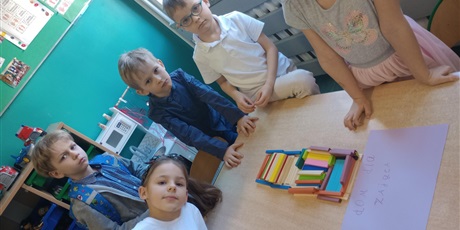 Powiększ grafikę: Dwie dziewczynki i trzech chłopców prezentuje swój dom dla zająca. Budowla wykonana jest z drewnianych klocków. 