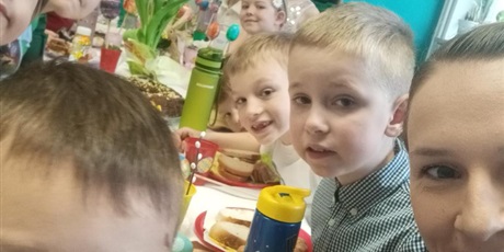 Powiększ grafikę: Grupowe selfie grupy 0A. Grupa siedzi przy stole. Na pierwszym planie widać chłopca w ciemnych włosach, panią nauczycielkę. Dzieci spoglądają w aparat. Na stole stoją żółte tulipany.