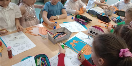 Powiększ grafikę: Grupa dzieci wykonuje pracę plastyczną "W wielkanocnym koszyczku". Na stole leżą kredki i kartki z pisankami i koszyczkami.
