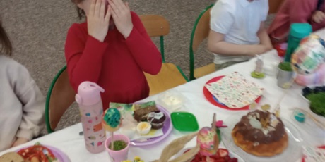 Powiększ grafikę: Śmigus dyngus. Dzieci siedzą przy stole i są opryskane wodą. Dziewczynka w czerwonej sukience zasłania oczy i twarz dłońmi.