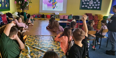 Powiększ grafikę: Grupa dzieci siedzi przy stole i i patrzy na ekran ścienny, na którym wyświetlona jest mapa Afryki. Jeden chłopiec ma uniesioną rękę do góry. Obok stoi nauczycielka zwrócona w stronę dzieci.