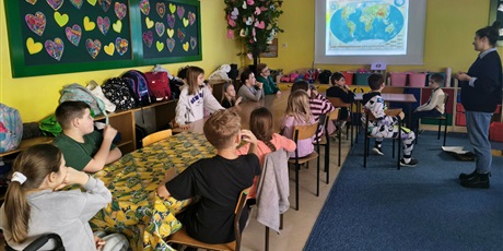 Powiększ grafikę: Grupa dzieci siedzi przy stole i i patrzy na ekran ścienny, na którym wyświetlona jest mapa świata. Obok stoi nauczycielka zwrócona w stronę dzieci.