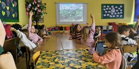 Powiększ grafikę: Grupa dzieci siedzi przy stole, kilkoro z nich ma uniesioną rękę do góry. W tle na ekranie ściennym wyświetlone jest zdjęcie lecących bocianów.