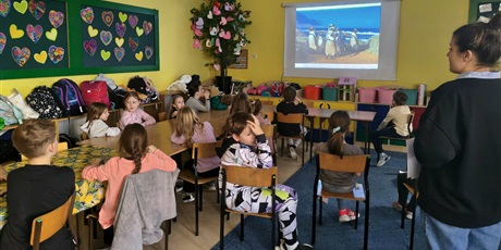 Powiększ grafikę: Grupa dzieci siedzi przy stole w świetlicy. Obok stoi nauczycielka zwrócona w stronę dzieci.