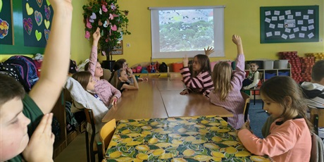 Powiększ grafikę: Grupa dzieci siedzi przy stole, kilkoro z nich ma uniesioną rękę do góry. W tle na ekranie ściennym wyświetlone jest zdjęcie lecących bocianów.