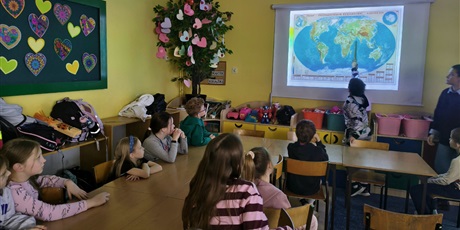 Powiększ grafikę:  Grupa dzieci siedzi przy stole zwrócona w stronę chłopca. Chłopiec pokazuje wskaźnikiem na mapie świata Afrykę. Obok stoi nauczycielka.