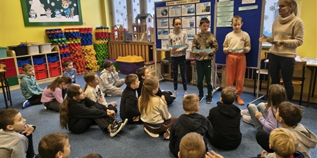 Powiększ grafikę: Grupa dzieci siedzi na dywanie zwrócona w kierunku tablic informacyjnych. Przy tablicach stoi troje uczniów i nauczycielka. Dzieci w rękach trzymają karteczki, z których czytają informacje o Antarktyd