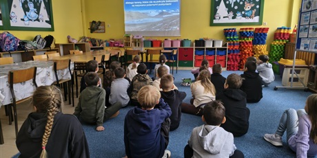 Powiększ grafikę: Grupa dzieci siedzi na dywanie tyłem do obiektywu. Uczniowi oglądają film wyświetlany na ściennym ekranie.