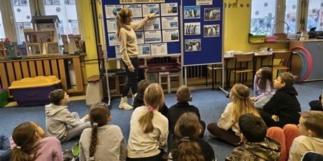 Powiększ grafikę: Nauczycielka stoi przy granatowych tablicach i prezentuje wiadomości o Antarktydzie. Przed nią na dywanie siedzą uczniowie, którzy w skupieniu słuchają.