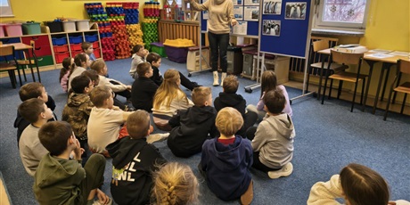 Powiększ grafikę: Nauczycielka stoi przy granatowych tablicach i prezentuje wiadomości o Antarktydzie. Przed nią na dywanie siedzą uczniowie, którzy w skupieniu słuchają.