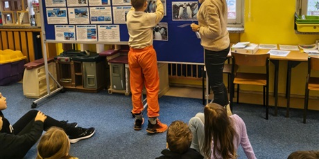 Powiększ grafikę: Chłopiec wiesza kartkę z nazwą pingwinów na granatową tablicę; obok stoi nauczycielka. Na dywanie siedzą dzieci i wpatrują się w tablicę.