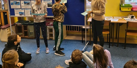 Powiększ grafikę: Przy tablicach stoi dwoje uczniów i nauczycielka. Dziewczynka w rękach trzyma kartkę, z której czyta informacje o Antarktydzie. Chłopiec wiesza kartkę na tablicy.