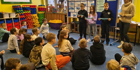 Powiększ grafikę: Grupa dzieci siedzi na dywanie zwrócona w kierunku tablic informacyjnych. Przy tablicach stoi troje uczniów i nauczycielka. Dzieci w rękach trzymają karteczki, z których czytają informacje o Antarktyd