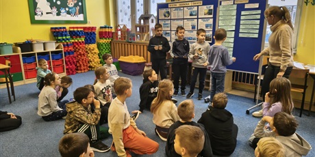 Powiększ grafikę: Grupa dzieci siedzi na dywanie zwrócona w kierunku tablic informacyjnych. Przy tablicach stoi czworo uczniów i nauczycielka. Dzieci trzymają karteczki, z których czytają informacje o Antarktydzie.