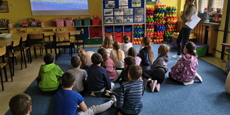 Powiększ grafikę: Grupa dzieci siedzi na dywanie w świetlicy tyłem do obiektywu. Obok stoi nauczycielka trzymająca białą kartkę. Wszyscy patrzą na film wyświetlany na ściennym ekranie.