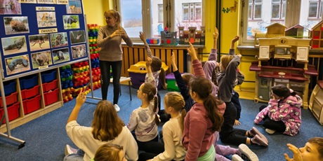 Powiększ grafikę: Grupa dzieci siedzi lub klęczy na dywanie. Część ma uniesioną rękę w górę. Wszyscy zwróceni są w stronę nauczycielki, która stoi obok granatowej tablica z napisem "W 8 dni dookoła świata".