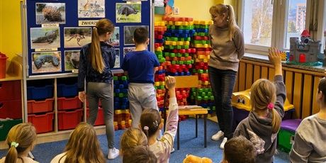 Powiększ grafikę: Dziewczynka i chłopiec zawieszają na granatowej tablicy napisy z nazwami australijskich zwierząt dopasowując je od zdjęć. Obok stoi nauczycielka. Kilkoro dzieci siedzi na dywanie i ma uniesiona rękę