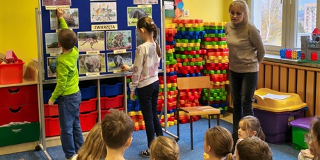 Powiększ grafikę: Dziewczynka i chłopiec zawieszają na granatowej tablicy napisy z nazwami australijskich zwierząt dopasowując je od zdjęć. Obok stoi nauczycielka, która mówi do dzieci siedzących na dywanie.