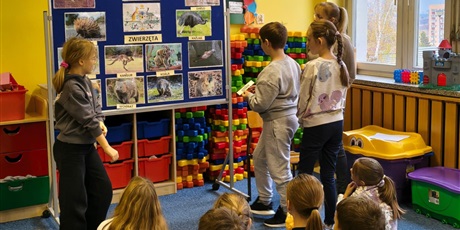 Powiększ grafikę: Dwie dziewczynki i chłopiec zawieszają na granatowej tablicy napisy z nazwami australijskich zwierząt dopasowując je od zdjęć. Obok stoi nauczycielka. Kilkoro dzieci siedzi na dywanie