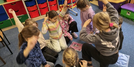 Powiększ grafikę: Dzieci siedzą na dywanie. Pięć dziewczynek ma uniesioną rękę do góry. Między nimi na podłodze leżą ułożone puzzle przedstawiające australijskie zwierzęta.