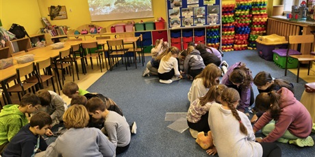 Powiększ grafikę: Trzy grupy dzieci siedzą na dywanie w świetlicy. Wszyscy pochylają się nad puzzlami przedstawiającymi australijskie zwierzęta. W tle znajduje się granatowa tablica z napisem "W 8 dni dookoła świata", 