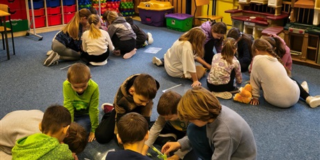 Powiększ grafikę: Trzy grupy dzieci siedzą na dywanie w świetlicy. Wszyscy pochylają się nad puzzlami przedstawiającymi australijskie zwierzęta. W tle znajduje się granatowa tablica z napisem "W 8 dni dookoła świata", 