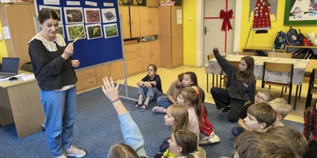 Powiększ grafikę: Grupa dzieci siedzi na dywanie w świetlicy i patrzy na nauczycielkę, która trzyma w ręku drewniane pałeczki. Kilkoro z nich ma uniesioną rękę do góry. W centralnym miejscu stoi granatowa tablica infor