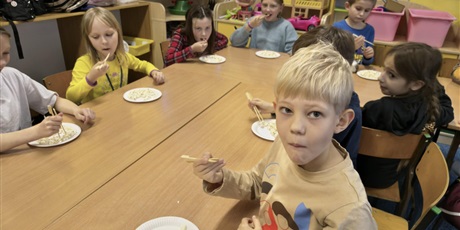 Powiększ grafikę: Grupa dzieci siedzi przy stole. Przed nimi stoją talerzyki z popcornem, który dzieci jedzą drewnianymi pałeczkami.