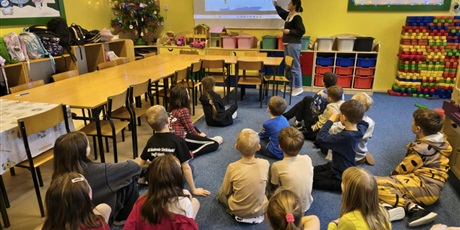 Powiększ grafikę: Grupa dzieci siedzi na dywanie w świetlicy patrząc na mapę wyświetlaną na ekranie ściennym. Nauczycielka opowiada i wskazuje ręką na kontynent.