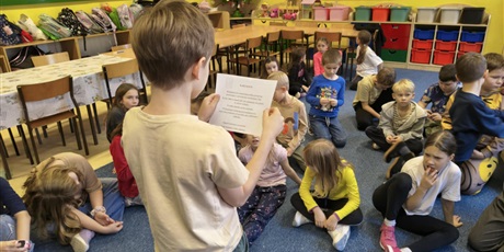 Powiększ grafikę: Chłopiec jasnej koszulce stoi zwrócony w stronę dzieci siedzących na dywanie. W ręku trzyma kartkę i czyta informację o przyprawach.