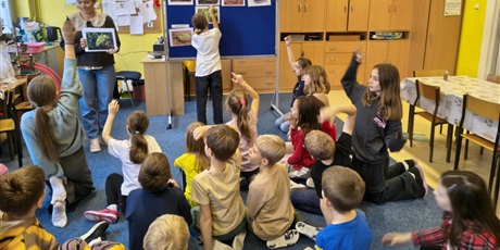 Powiększ grafikę: Grupa dzieci siedzi na dywanie w świetlicy i patrzy na nauczycielkę. Kilkoro z nich ma uniesioną rękę do góry. W centralnym miejscu stoi granatowa tablica informacyjna, na ktorej dziewczynka wiesza il