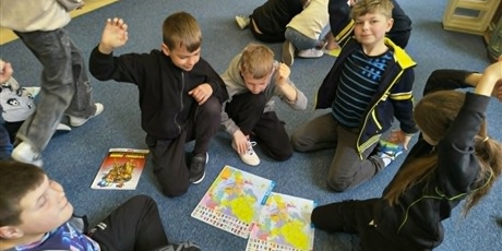 Powiększ grafikę: Grupa dzieci siedzi na dywanie i podnosi ręce do góry. Przed nimi leżą ułożone puzzle przedstawiające mapę Europy.