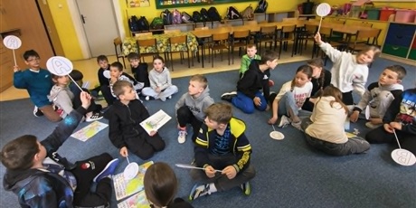 Powiększ grafikę: Dzieci siedzą na dywanie. Troje z nich ma uniesione do góry patyki z literką oznaczającą wybór odpowiedzi.