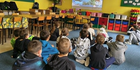 Powiększ grafikę: Grupa dzieci siedzi na dywanie tyłem do obiektywu. Uczniowi oglądają film wyświetlany na ściennym ekranie.