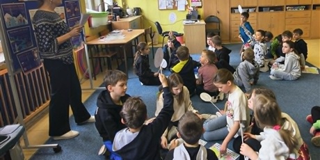 Powiększ grafikę: Nauczycielka stoi przy granatowej tablicy.  Dzieci siedzą na dywanie. Niektóre z nich mają uniesione do góry patyki z literką oznaczającą wybór odpowiedzi.