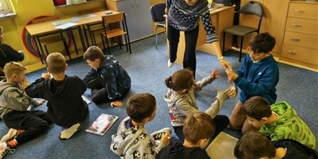 Powiększ grafikę: Dzieci siedzą na dywanie i układają puzzle. Nauczycielka podaje uczniom woreczek z brakującymi puzzlami.