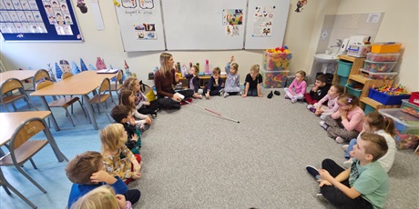 Powiększ grafikę: Grupa 6-letnich dzieci siedzi na dywanie w kole. Słuchają tego co mówi prowadząca, która pokazuje dzieciom biała laskę. 