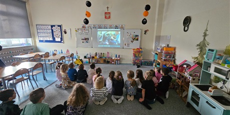 Powiększ grafikę: Dzieci siedząc na dywanie oglądają prezentację przygotowaną przez prowadzącą.